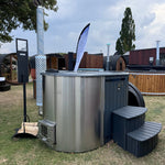 A wood-fired ofuro style hot tub with integrated heater, featuring a fibreglass liner and surrounded by wooden steps, a chimney guard, and a drinks shelf.