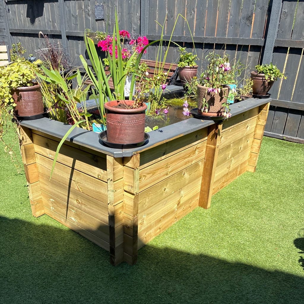 A wooden raised garden pond with a black liner and potted plants, set on artificial grass in a backyard.