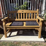 A wooden outdoor bench with two dark gray seat cushions, placed on a concrete slab in a backyard setting with grass, plants, and a white railing behind it.