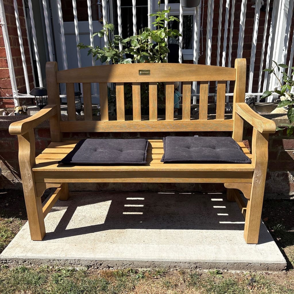 A wooden outdoor bench with two dark gray seat cushions, placed on a concrete slab in a backyard setting with grass, plants, and a white railing behind it.