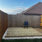 A wooden deck platform in a backyard, bordered by white gravel and green grass, with a tall wooden fence behind it and a blue folding chair placed on the deck. A house roof is visible in the background under a blue sky.