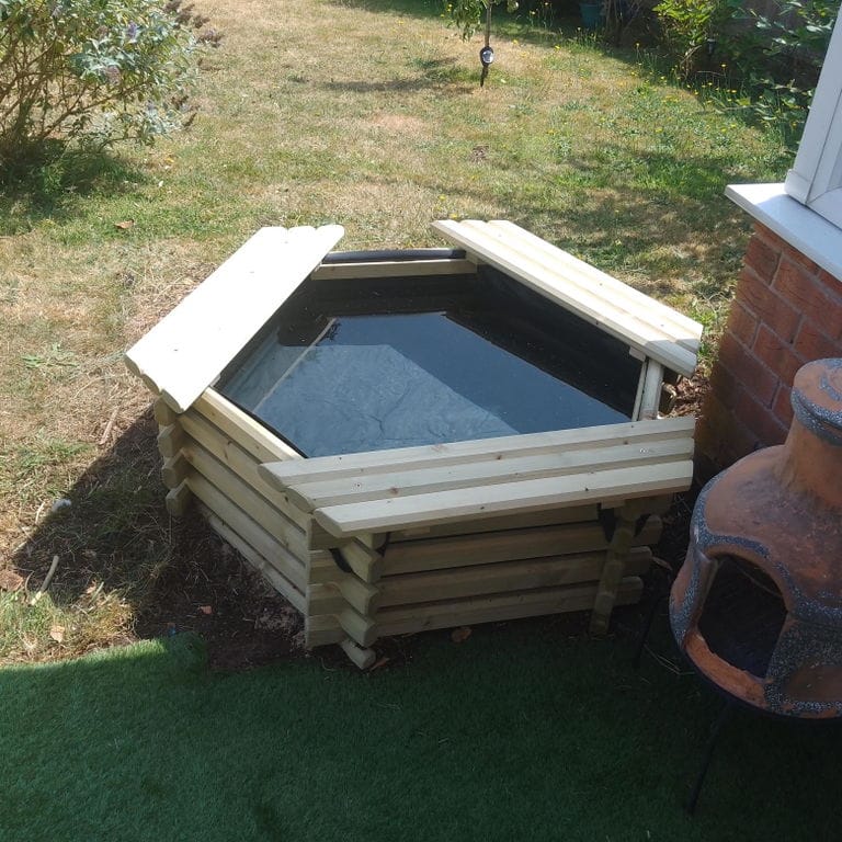 A hexagonal wooden outdoor pond with a black liner, shown in a backyard next to a brick chimney and artificial grass.