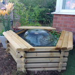 A wooden raised garden bed with a glass dome planter containing water and greenery, set in a backyard garden next to a brick wall and potted lilies.