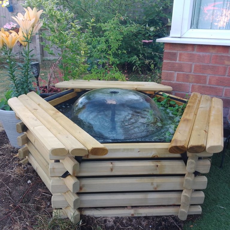 A wooden raised garden bed with a glass dome planter containing water and greenery, set in a backyard garden next to a brick wall and potted lilies.