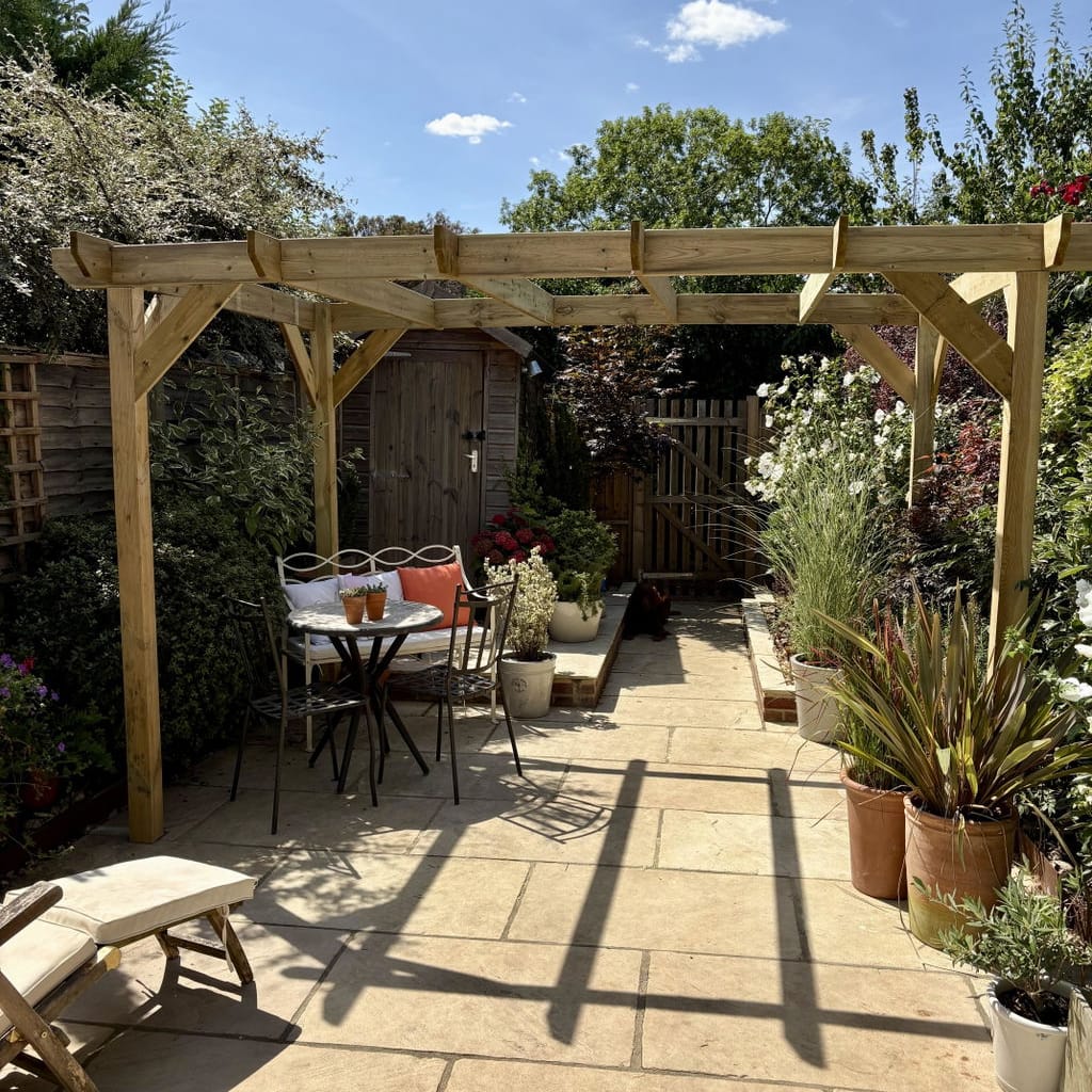 A wooden garden pergola with a dining table and chairs underneath, set in a landscaped backyard with potted plants, a stone patio, and a blue sky background.