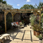 A wooden garden pergola with a dining table and chairs underneath, set in a landscaped backyard with potted plants, a stone patio, and a blue sky background.
