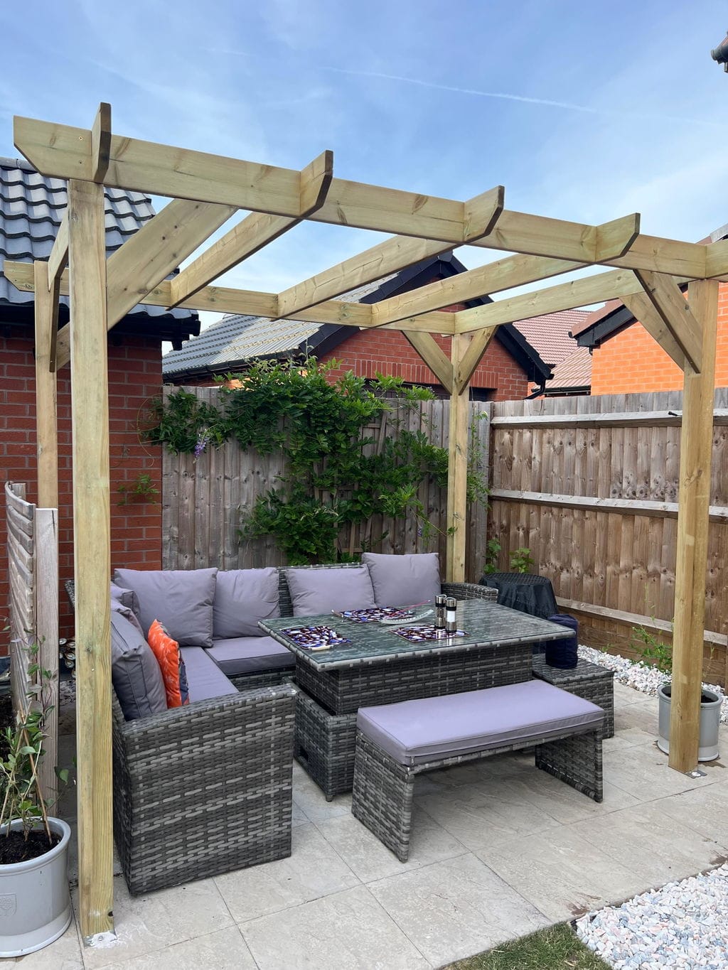 Outdoor patio furniture set under a wooden pergola in a backyard, including a gray wicker sectional sofa with light purple cushions, a matching rectangular coffee table, and a long bench, surrounded by a wooden fence and brick house.