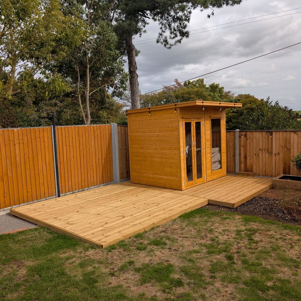A small wooden garden shed with double glass doors sits on a raised wooden deck in a backyard with grass, trees, and a wooden fence.