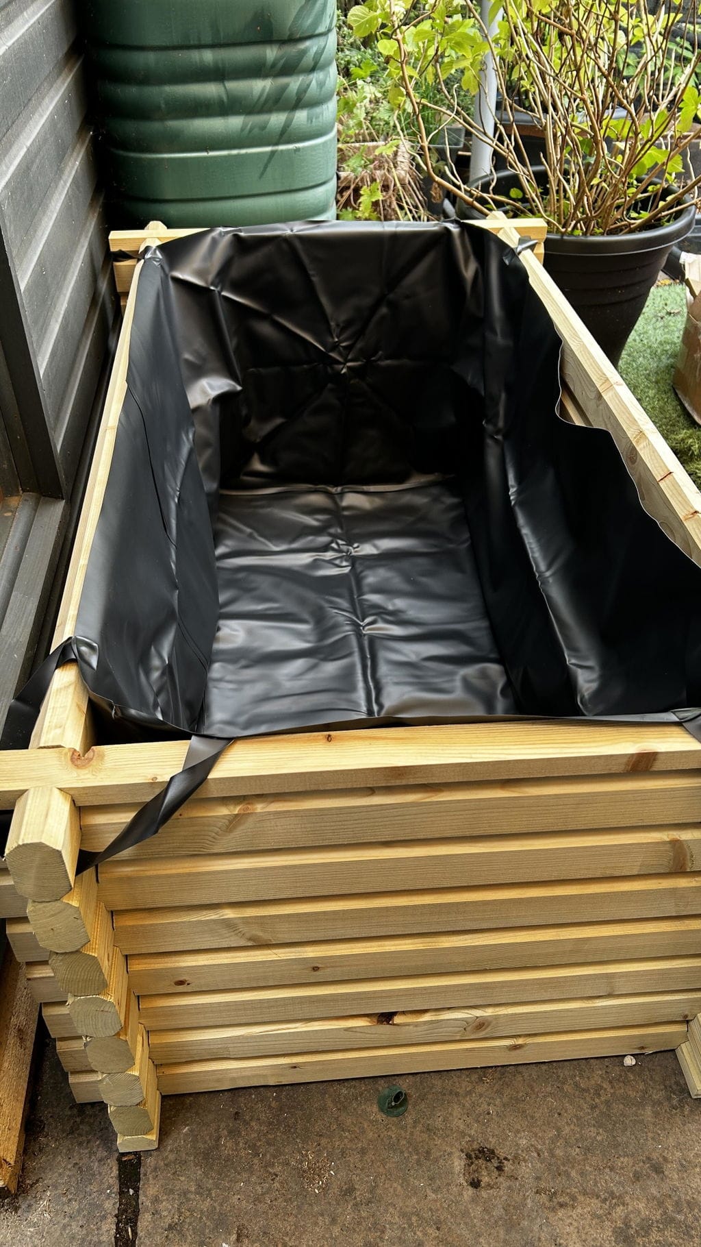 A wooden raised garden pond with a black waterproof liner inside, photographed outdoors next to a green barrel and potted plants.
