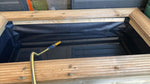 A wooden-framed outdoor pond liner or fish pond with a black liner inside, shown with a yellow hose connected near the bottom.