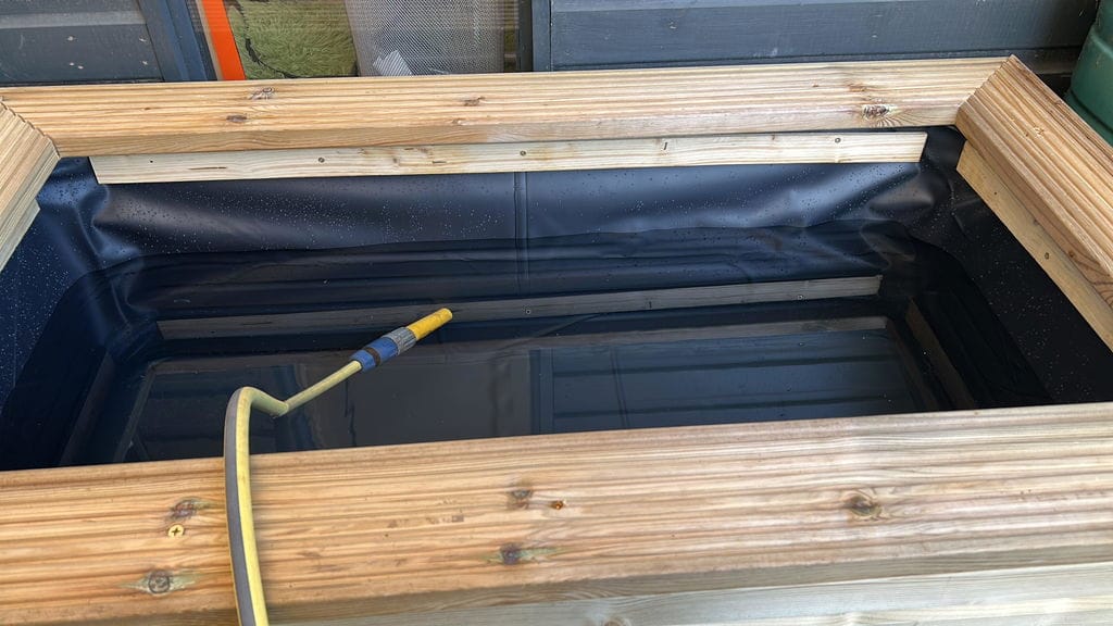 A wooden-framed outdoor pond liner or fish pond with a black liner inside, shown with a yellow hose connected near the bottom.