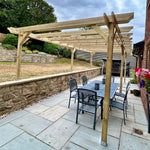 A wooden pergola structure installed on a paved backyard patio, featuring a dining table and chairs underneath. The patio is bordered by a stone retaining wall on one side and a red brick house wall with flower boxes on the other, with a grassy lawn and trees in the background under a cloudy sky.