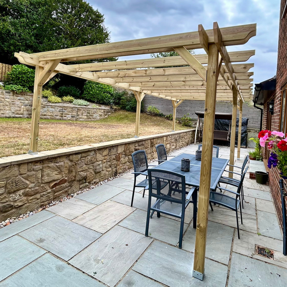 A wooden pergola structure installed on a paved backyard patio, featuring a dining table and chairs underneath. The patio is bordered by a stone retaining wall on one side and a red brick house wall with flower boxes on the other, with a grassy lawn and trees in the background under a cloudy sky.