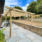 A wooden pergola with a slatted roof extends over a paved patio area with outdoor dining furniture, bordered by a stone retaining wall that separates the patio from a grassy backyard with trees and a fence under a blue sky.