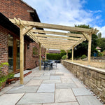 A wooden pergola structure installed over a paved patio in a backyard setting next to a brick house, with outdoor dining furniture, a stone wall, and a blue sky with clouds.