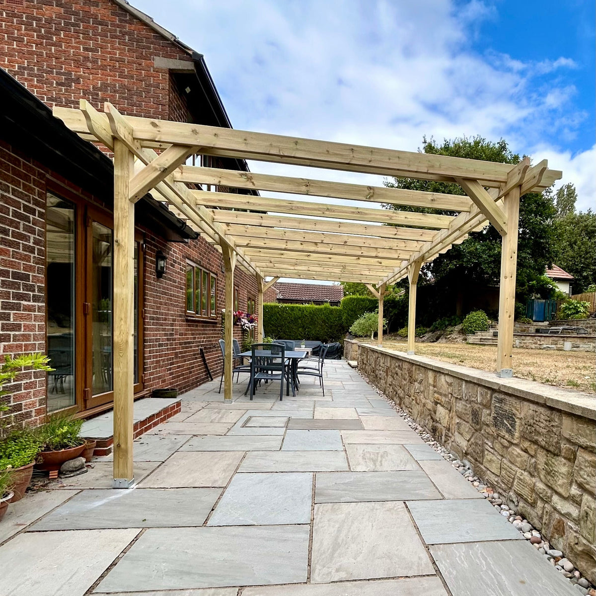 A wooden pergola structure installed over a paved patio in a backyard setting next to a brick house, with outdoor dining furniture, a stone wall, and a blue sky with clouds.