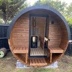A round wooden sauna cabin with a dark gray curved exterior shell, open front door, and built-in wooden benches, photographed outdoors on grass with trees and a fence in the background.