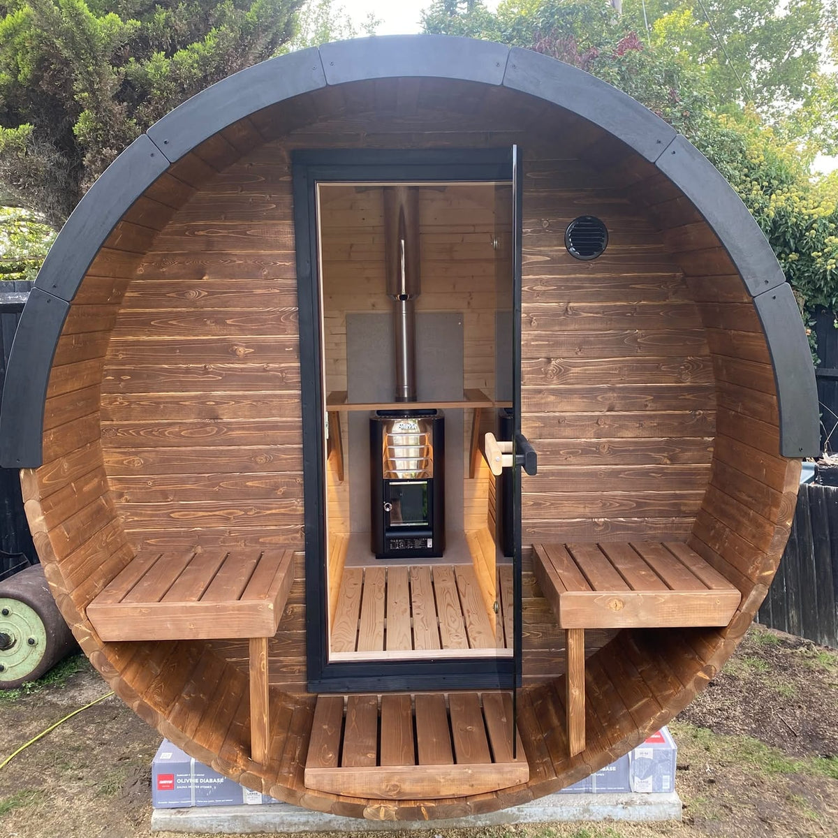 A round wooden sauna cabin with a black metal frame and door, featuring built-in wooden benches and a central wood-burning stove, photographed outdoors on grass with trees in the background.
