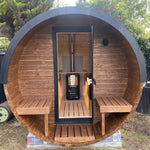 A round wooden sauna cabin with a black metal frame and door, featuring built-in wooden benches and a central wood-burning stove, photographed outdoors on grass with trees in the background.
