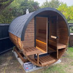 A round wooden sauna cabin with a dark gray shingled roof, glass door, and built-in wooden benches, shown outdoors on a grassy lawn with trees and a fence in the background.