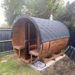 A wooden barrel-style sauna cabin with a dark gray shingled dome roof, set outdoors on a concrete base in a backyard with grass, trees, and a fence visible.