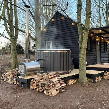 A large wood-fired hot tub with an external heater, featuring a stainless steel chimney and a faux leather lid, set on a wooden surface.